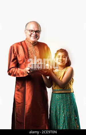 Homme âgé ou âgé indien avec petite-fille célébrant son anniversaire en soufflant des bougies sur un gâteau tout en portant des vêtements ethniques, se tenant isolé du blanc Banque D'Images