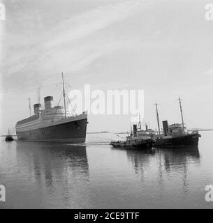 Dans les années 1950, les remorqueurs historiques tirent le navire de mer RMS Queen Elizabeth dans les eaux calmes du port français de Cherbourg. Nommé d'après la reine mère britannique, il était le plus grand paquebot jamais construit à cette époque (1938) et est resté ainsi pendant 56 ans par la suite. Elle a été utilisée comme navire de guerre pendant la Seconde Guerre mondiale et n'est devenue qu'un paquebot, comme prévu, en 1946. À partir de là, ce paquebot de luxe a assuré un service hebdomadaire entre Southampton, Royaume-Uni et New York, aux États-Unis, via Cherbourg, France. En 1972, dans le port de Victoria, à Hong Kong, lors d'une remise en état, elle a pris le feu et a coulé. Banque D'Images