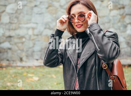 Femme souriante habillée style boho de mode dans une veste de motard en cuir noir avec sac à rabat en cuir marron ayant une marche de parc d'automne. Banque D'Images