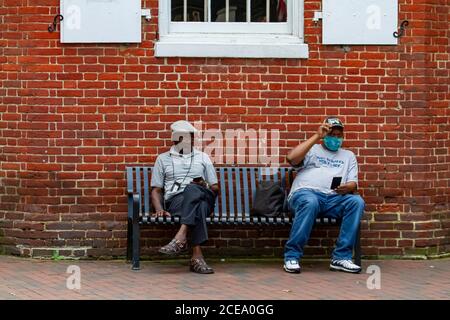 Annapolis, MD 08/21/2020: Deux hommes afro-américains âgés sont assis sur un banc à l'extérieur d'un vieux bâtiment en briques à Annapolis. L'un porte le masque facial dû Banque D'Images