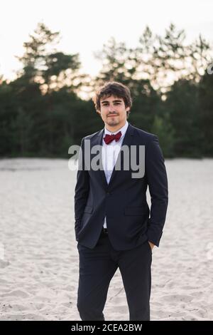 Homme adulte portant un élégant costume noir avec noeud papillon et se tenir sur la plage avec les mains dans les poches en regardant l'appareil photo Banque D'Images