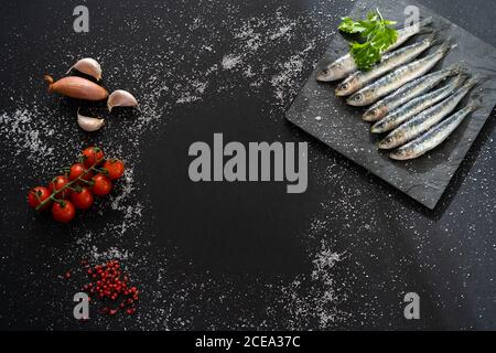 Tableau carré avec ensemble d'anchois frais couché sur du noir arrière-plan près de diverses épices et bouquet de tomates cerises mûres Banque D'Images