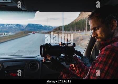 Homme barbu dans une chemise à carreaux assis sur le siège passager de la voiture et prenant des photos lors de votre voyage à travers l'Islande. Banque D'Images