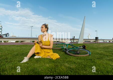 Femme élégante en robe jaune fluide et baskets assis sur la pelouse avec le vélo couché derrière et écrire avec un stylo dans un petit carnet qui regarde loin Banque D'Images