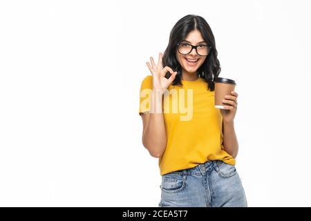 Jeune femme buvant une tasse de café à emporter sur fond blanc faisant signe ok avec les doigts, symbole excellent Banque D'Images