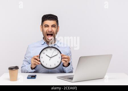 Gestion du temps. Furieuse a stressé un employé assis sur son lieu de travail avec un ordinateur portable, tenant une horloge et criant déprimé dans le bureau, en regardant fou Ove Banque D'Images