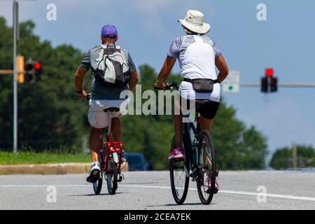Un couple senior portant des shorts et t-shirts décontractés ainsi que des chapeaux est à vélo près d'une autoroute par une belle journée d'été. L'homme a une moto à pédale pliable Banque D'Images