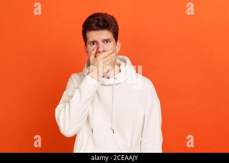 Oh mon Dieu, incroyable! Un jeune homme choqué avec une coiffure élégante dans un sweat à capuche décontracté fermant la bouche avec la main, en gardant des informations secrètes. Prise de vue en studio Banque D'Images