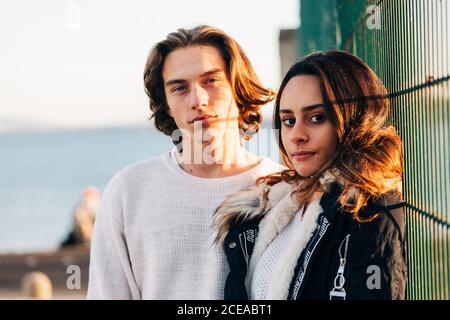 Homme heureux et belle femme heureuse en manteau regardant la caméra près de clôture en métal Banque D'Images