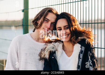 Homme heureux et belle femme heureuse en manteau regardant la caméra près de clôture en métal Banque D'Images