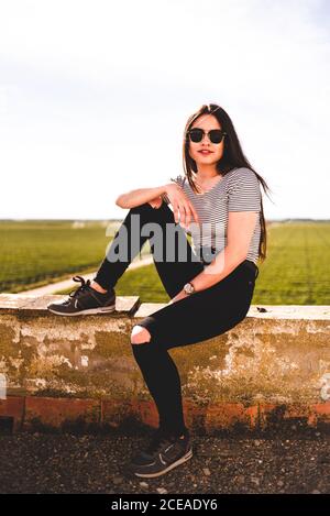Belle jeune femme en lunettes de soleil appréciant la journée ensoleillée tout en passant du temps dans le champ vert. Banque D'Images