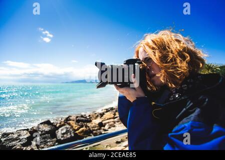 Vue latérale d'une jolie femme à tête rouge qui prend des photos de paysages marins sur la côte rocheuse par temps ensoleillé. Banque D'Images