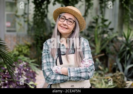 Gros plan sur une jeune femme gardenier souriante, avec de longs cheveux gris, portant un chapeau de paille et des vêtements de travail décontractés, posant à l'appareil photo avec les bras croisés Banque D'Images