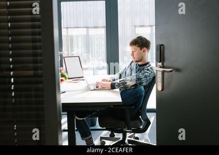 Vue latérale d'un homme dans des vêtements décontractés confortablement installés chaise de bureau et clavier pour ordinateur portable sur bureau blanc avec une grande fenêtre sur l'arrière-plan prise par la porte ouverte Banque D'Images