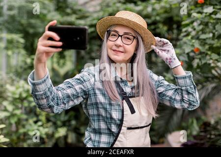 Portrait d'une jolie femme joyeuse des années 60 gardenier avec de longs cheveux gris, portant une chemise à carreaux, un tablier et un chapeau de paille, debout dans une belle orangerie Banque D'Images