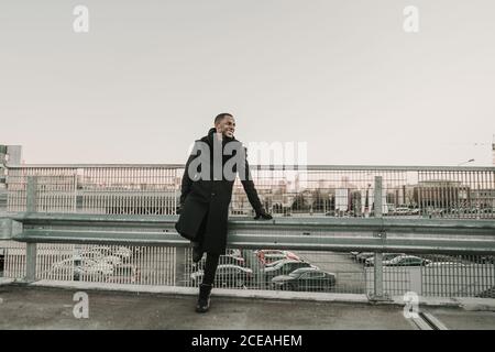 Beau homme afro-américain en manteau élégant souriant et regardant s'éloigner en s'inclinant sur la rambarde de dépassement en arrière-plan du stationnement lot et ville moderne Banque D'Images