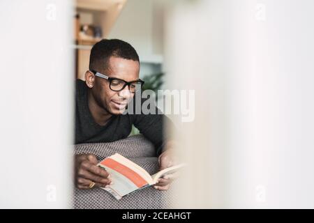 Homme afro-américain adulte en lunettes lisant le livre tout en étant assis sur le canapé à la maison Banque D'Images