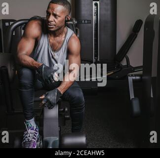 Séduisant Afro-américain gars dans sportswear à l'écoute de la musique dans ecouteurs et regarder loin en étant assis sur la machine d'exercice salle de sport moderne Banque D'Images