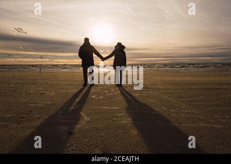 Vue de couple en extérieur sur la plage de sable à la saison froide tient les mains dans le dos éclairé Banque D'Images