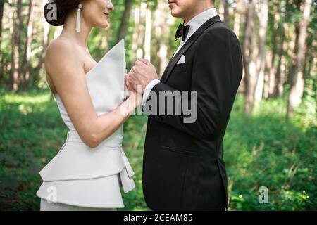 Vue latérale d'un jeune marié méconnu et d'une jeune mariée qui se touche les mains de l'autre tout en se tenant sur un arrière-plan flou de belle forêt le jour ensoleillé Banque D'Images