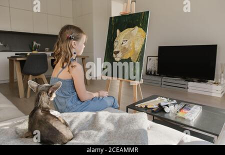 Vue latérale d'une petite fille talentueuse qui dessine des tableaux chevalet contre la fenêtre à la maison Banque D'Images