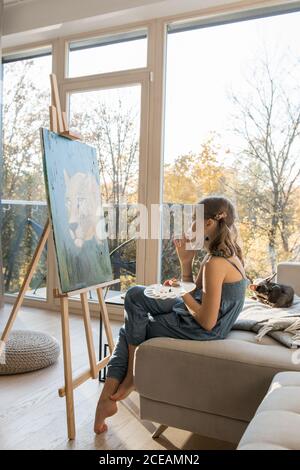 Vue latérale d'une petite fille talentueuse qui dessine des tableaux chevalet contre la fenêtre à la maison Banque D'Images