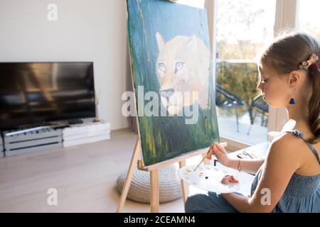 Vue latérale d'une petite fille talentueuse qui dessine des tableaux chevalet contre la fenêtre à la maison Banque D'Images