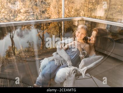 Tiré à travers la fenêtre en verre de la femme avec la petite fille et le chien en cuddling à carreaux tout en étant assis sur un fauteuil sur la terrasse Banque D'Images