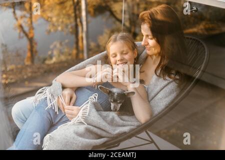 Tiré à travers la fenêtre en verre de la femme avec la petite fille et le chien en cuddling à carreaux tout en étant assis sur un fauteuil sur la terrasse Banque D'Images