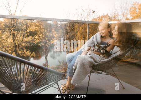 Tiré à travers la fenêtre en verre de la femme avec la petite fille et le chien en cuddling à carreaux tout en étant assis sur un fauteuil sur la terrasse Banque D'Images