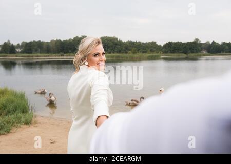 Adulte belle et élégante mariée tenant petit main de marié et sourire à la caméra tout en se tenant sur la côte du lac avec cygnes Banque D'Images