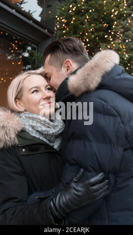 Vue latérale d'heureux guy hugging jeune femme vestes de ski près de l'arbre de Noël décoré par allumé fairy lights on city street Banque D'Images