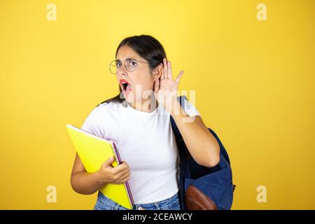 Jeune belle étudiante femme portant un sac à dos tenant un ordinateur portable sur isolé fond jaune surpris par l'écoute de la main sur l'oreille à r Banque D'Images