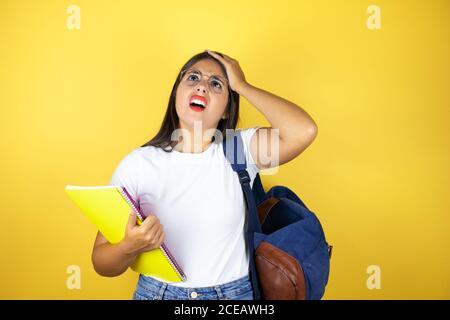 Jeune belle étudiante femme portant un sac à dos tenant un ordinateur portable sur isolé fond jaune mettant une main sur sa tête en souriant comme elle avait forg Banque D'Images