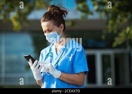 pandémie covid-19. triste médecin moderne femme dans des exfoliations avec stéthoscope et masque médical utilisant smartphone application à l'extérieur près de la clinique. Banque D'Images