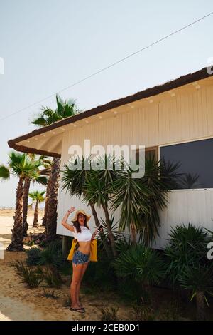 Femme amusante vêtue d'un t-shirt blanc et d'un short jean avec panama tenant sac jaune sur fond avec maison de plage et palmiers verts Banque D'Images