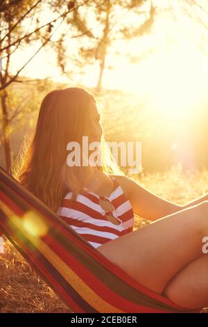 Charmante femme aux longs cheveux bruns vêtue de rouge et blanc maillot de bain nu allongé dans un hamac coloré sur fond de soleil lumineux et de feuillage Banque D'Images