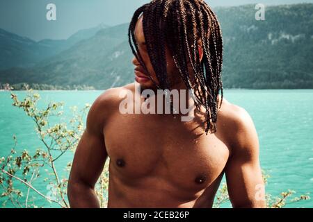 Vue rognée d'un jeune homme athlétique afro-américain élégant qui regarde loin Près d'un magnifique lac entre les montagnes en Autriche Banque D'Images