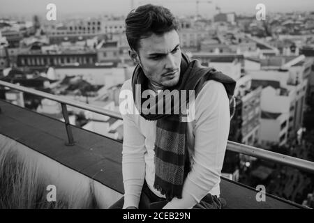 Noir et blanc jeune homme élégant en tenue décontractée avec foulard assis sur le toit et regardant loin sur fond urbain à Madrid, Espagne Banque D'Images