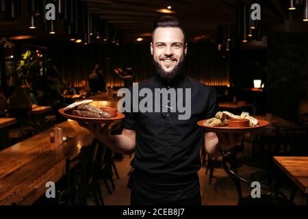 Serveur à la barbe gaie avec kebab et accompagnement dans un restaurant géorgien. Un serveur éclairé en vêtements noirs, avec une barbe et une portion de kebabs Banque D'Images