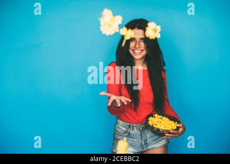 Jeune femme attrayante avec de longs cheveux foncés dans un chemisier rouge et un short en denim debout en studio et jetant des fleurs jaunes dans l'air sur fond bleu Banque D'Images