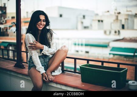 Jeune belle femme avec de longs cheveux foncés assis au balcon et souriant sur fond de maisons Banque D'Images