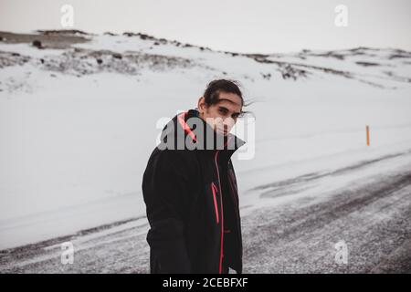 Vue latérale d'un beau gars dans des vêtements chauds regardant caméra sur une route de campagne enneigée en hiver venteux Journée en Islande Banque D'Images