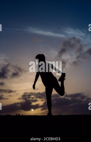 Vue arrière de silhouette de jeune femme qui s'étend les jambes dans les vêtements de sport et de merveilleux ciel avec nuages en soirée Banque D'Images