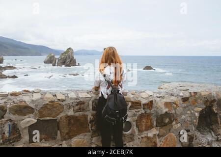 Vue arrière de la jeune femme avec sac à dos debout près du vieux mur de pierre et regardant la belle mer sur l'îlot de Gatzelugatxe en Espagne Banque D'Images