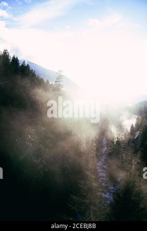 Vue en perspective du brouillard sous la lumière du soleil au-dessus de la vallée rocheuse enneigée avec ruisseau coulant parmi les conifères. Banque D'Images