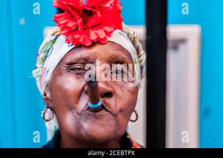 CUBA - 14 septembre 2017 : vue rapprochée de la jeune femme afro-cubaine senior avec une grande fleur rouge sur la bruyère tenant le cigare dans la bouche et regardant l'appareil photo sur un arrière-plan flou Banque D'Images