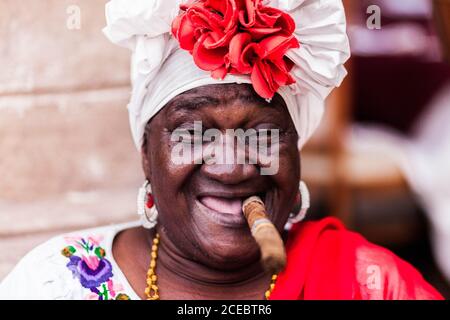 CUBA - 14 septembre 2017 : vue rapprochée de la jeune femme afro-cubaine senior avec une grande fleur rouge sur la bruyère tenant le cigare dans la bouche et regardant l'appareil photo sur un arrière-plan flou Banque D'Images