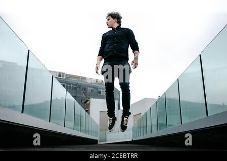 Jeune homme tatoué dans des vêtements noirs sautant facilement sur la marche pont avec clôture en verre et vue sur les bâtiments arrière-plan Banque D'Images