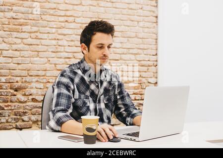 Homme concentré travaillant avec un ordinateur portable et assis à table avec mug et téléphone portable près d'un mur de briques Banque D'Images